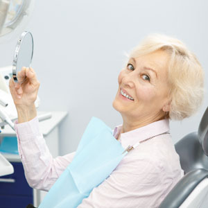 Senior female patient smiling to the camera holding a mirror sitting in a dental chair at the clinic